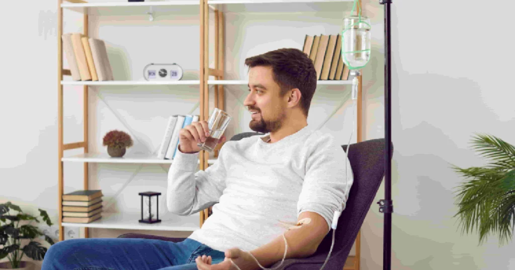 A smiling man sits in a chair with an IV drip in his arm, holding a glass of water during his IV Therapy in Glenwood Springs, CO.
