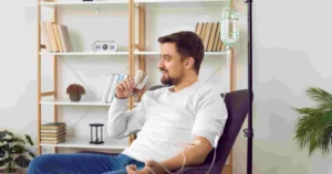 A smiling man sits in a chair with an IV drip in his arm, holding a glass of water during his IV Therapy in Glenwood Springs, CO.