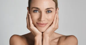 A close-up portrait of a woman with blue eyes framing her face with her hands, showcasing BBL HEROiC in Glenwood, against a grey background.